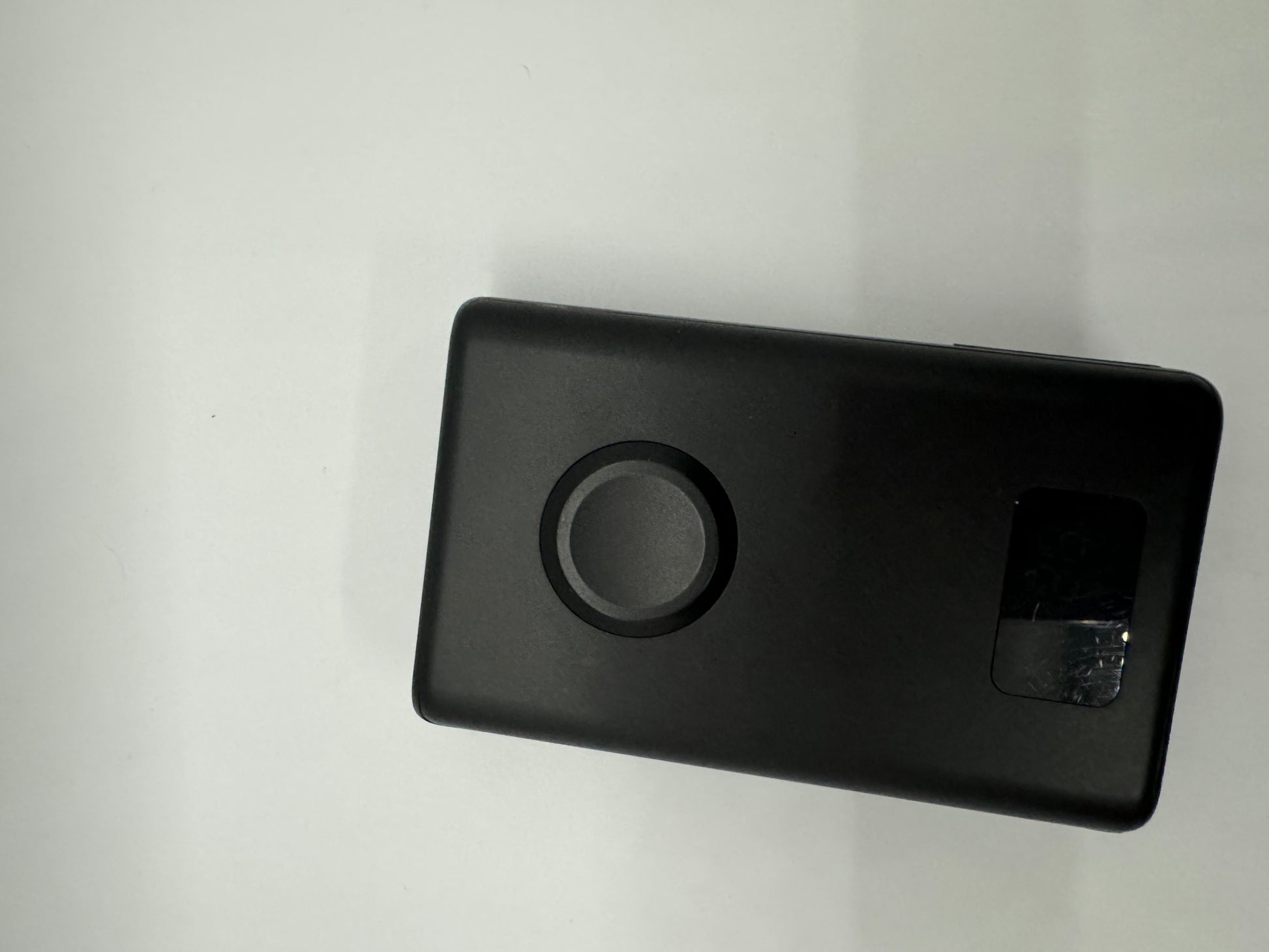 The picture shows a black rectangular object with rounded corners on a white background. The object is positioned in the bottom right corner of the image. On the object, there is a circular indentation or button near the center, and a small square cut-out on the right side. The square cut-out seems to have something reflective or metallic inside. The surface of the object appears to be smooth and matte.