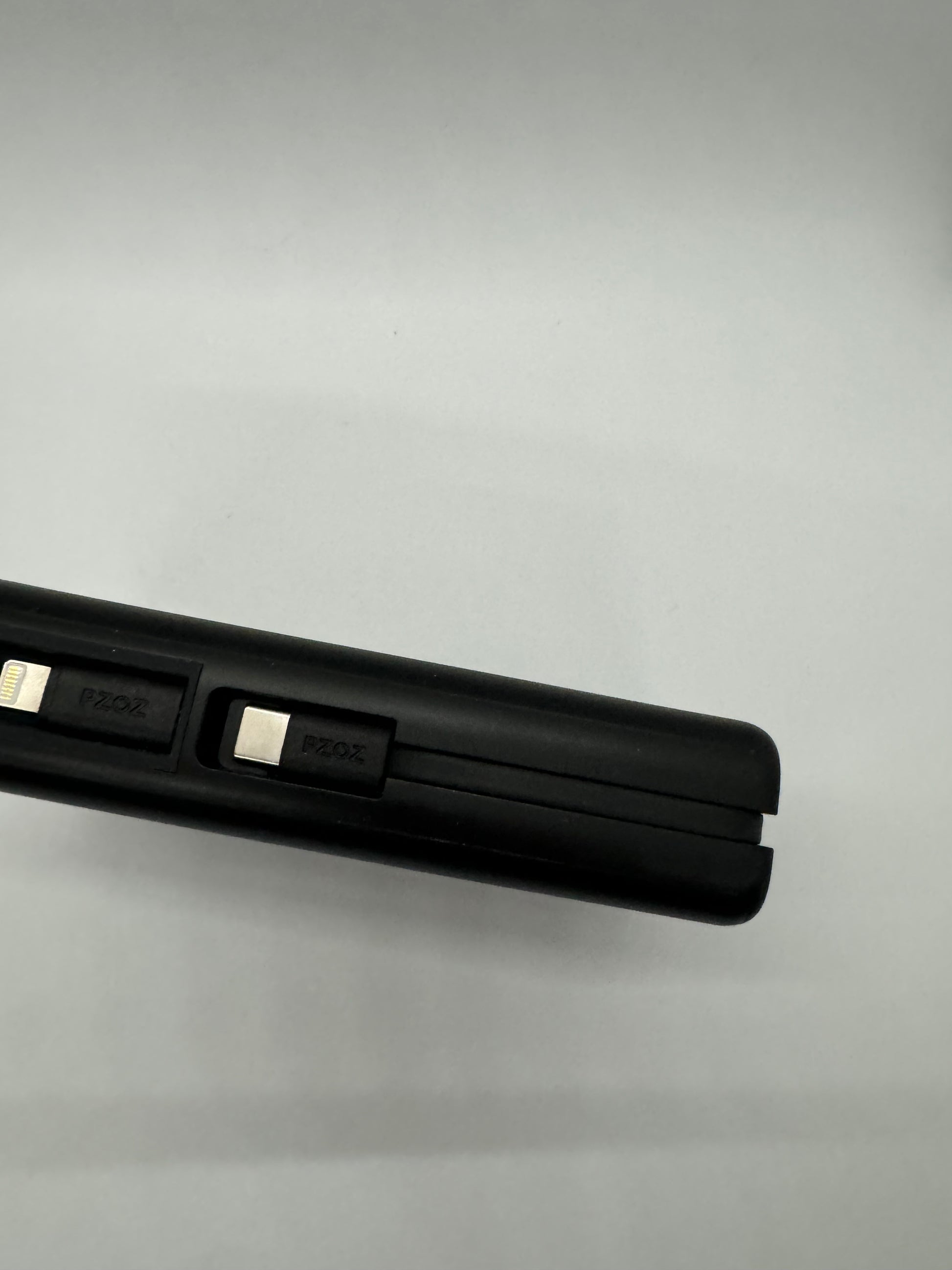 The picture shows a black object in the bottom right corner against a plain white background. The object appears to be a slim, rectangular device with rounded edges. It has two slots on the top side, each labeled "PZOZ". There is also a small, rectangular metallic connector protruding from the left side of the device. The device seems to be some sort of electronic accessory, possibly a charging or connection cable.