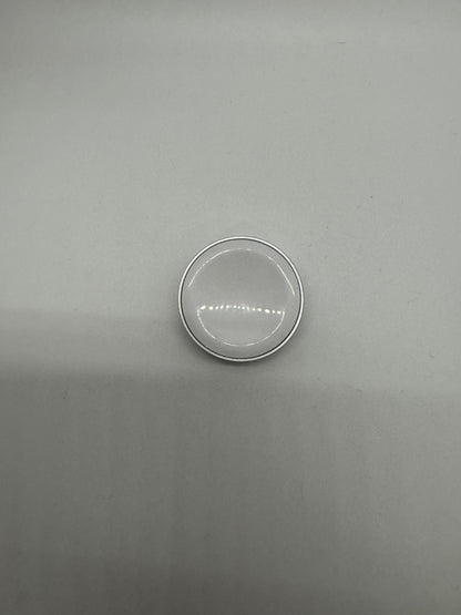 The picture shows a small, circular object placed on a plain surface. The object appears to be a silver-colored metal ring with a transparent center. The transparent center has a horizontal pattern of lines. The background is a plain, light-colored surface. The object is positioned towards the bottom of the image.