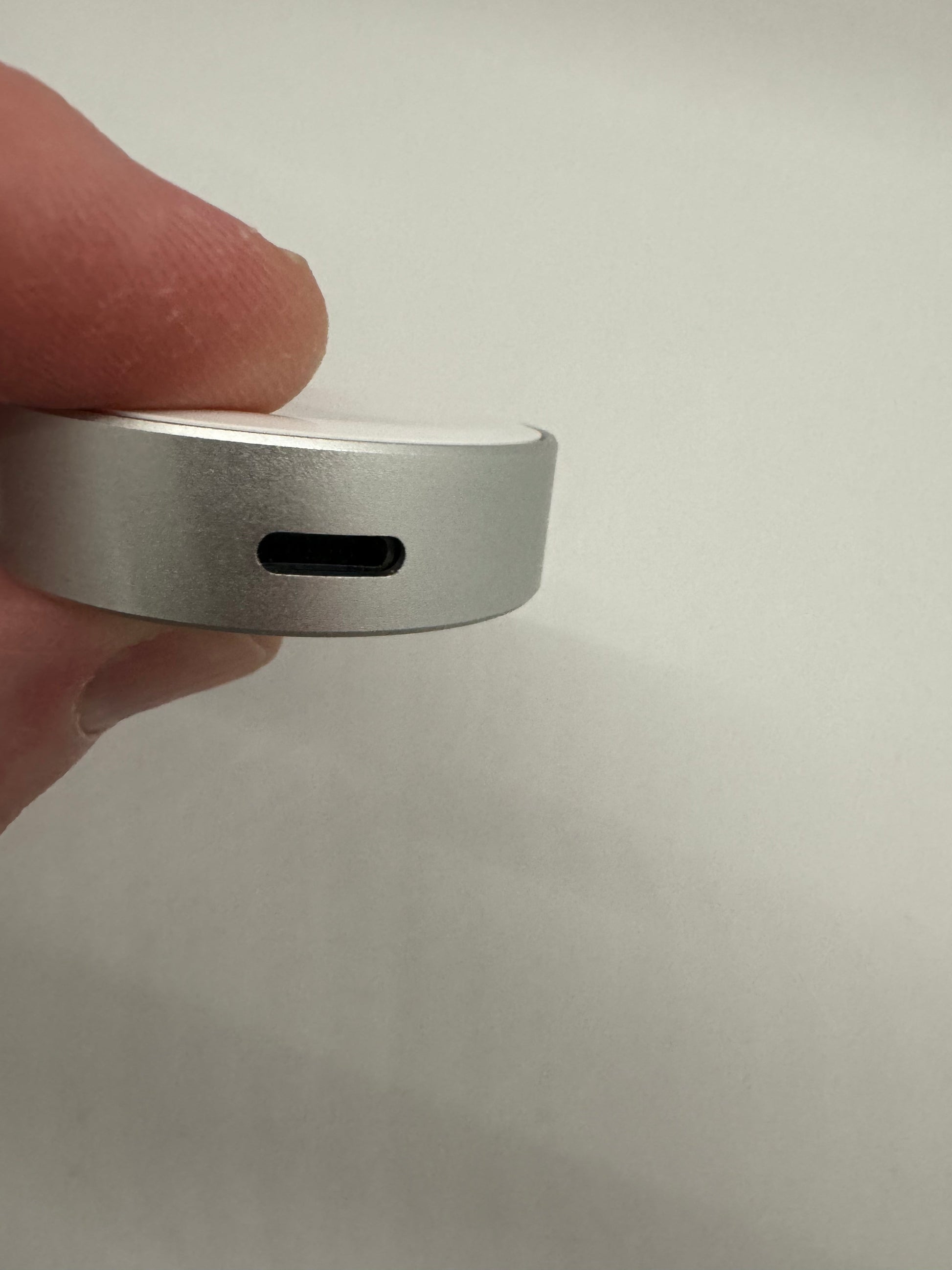 Be My AI: The picture shows a close-up of a person's fingers holding a small, silver, circular device. The device appears to be made of metal and has a single rectangular port on its side. The background is plain and white.