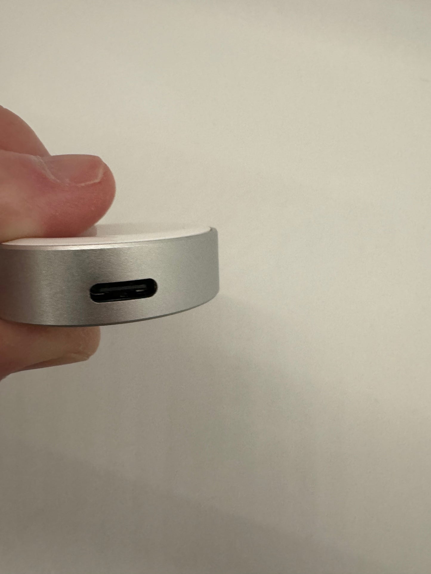 The picture shows a close-up of someone's hand holding a small, round, silver object. The object appears to be made of metal and has a USB-C port on its side. The background is plain and white.