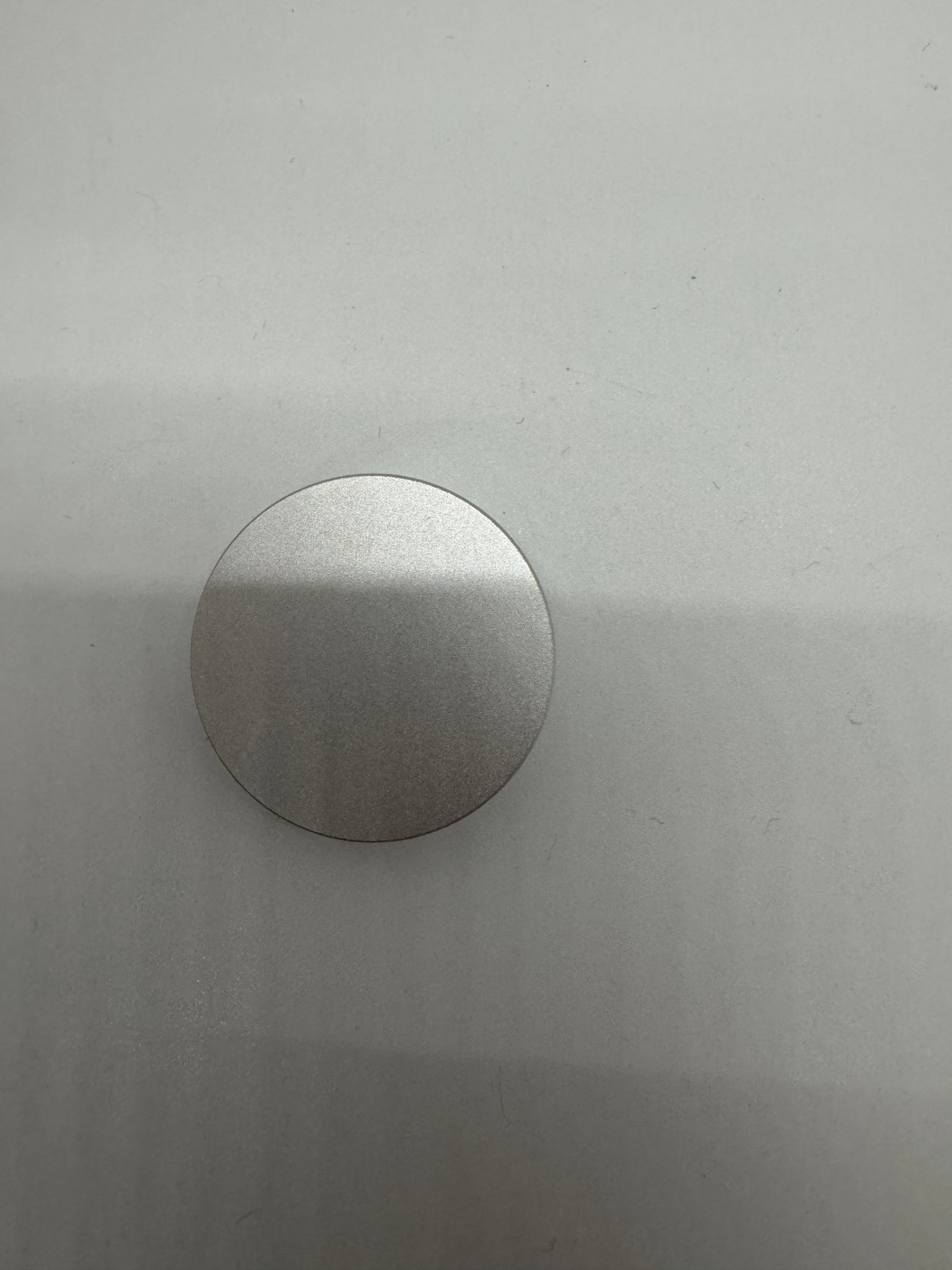 The picture shows a silver, circular object on a light grey background. The object appears to be metallic and flat. It is positioned towards the bottom of the image, and only the top half of the circle is visible. The background has a smooth texture with some minor scratches and specks.