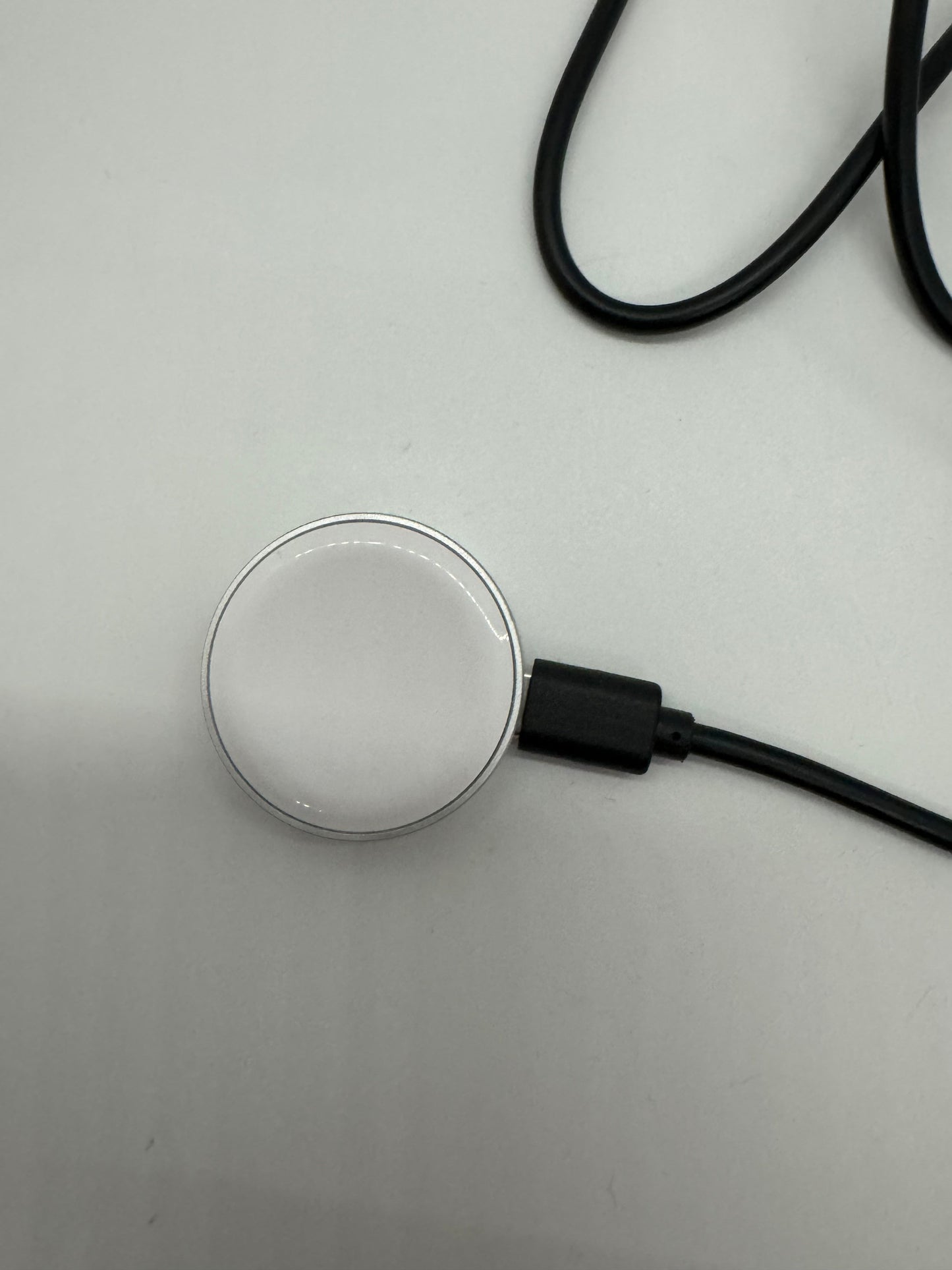 The picture shows a round, silver object that appears to be a wireless charger. It is placed on a white surface. There is a black cable attached to it, which has a black rectangular attachment near where the cable connects to the round object. The cable is coiled in the upper part of the picture.
