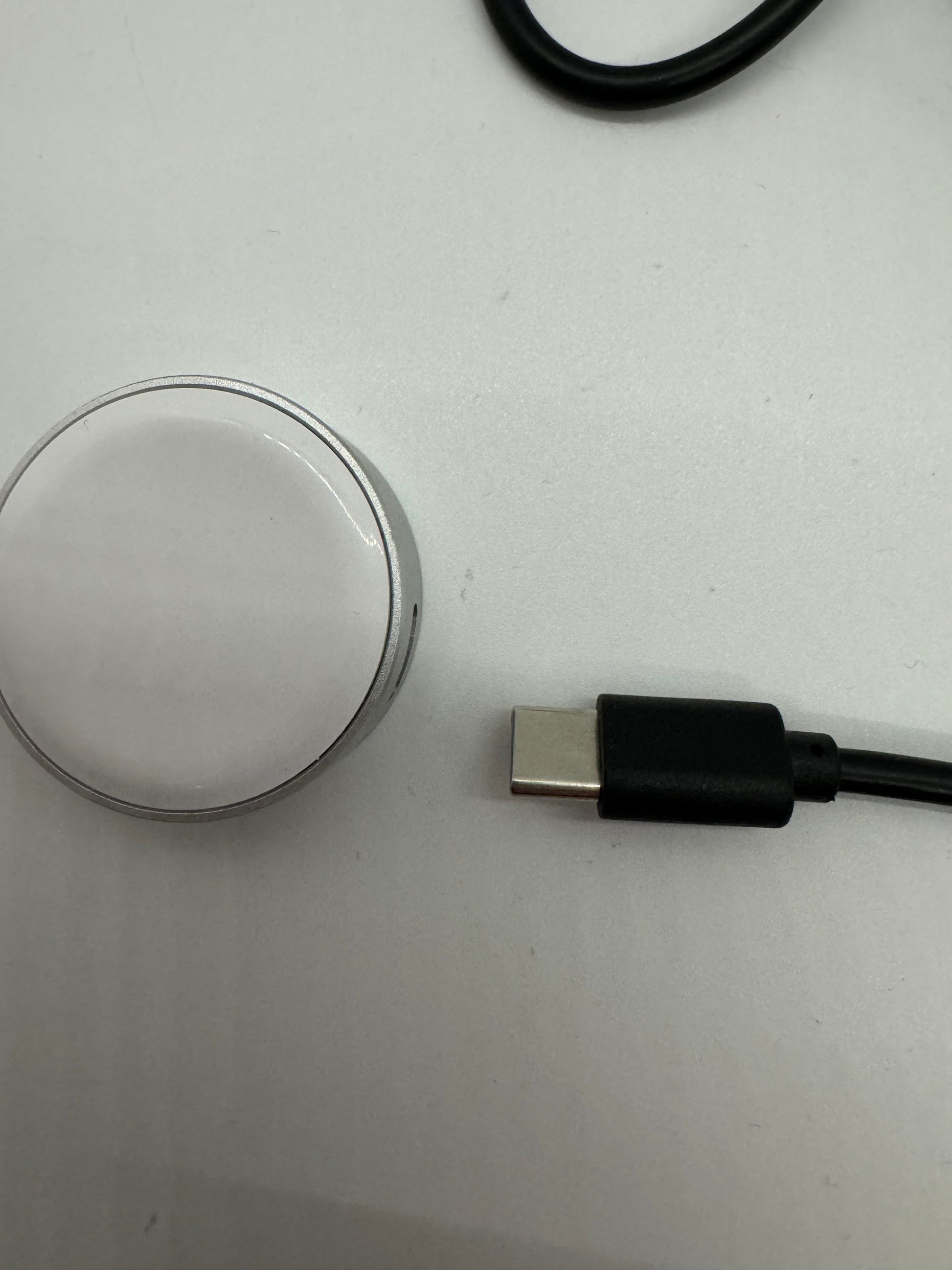 The picture shows two objects on a white surface. On the left side, there is a round object that appears to be a small mirror or a metal disk. It has a reflective surface and a thin metal edge. On the right side, there is a black cable with a USB connector at the end. The USB connector is silver and rectangular in shape.