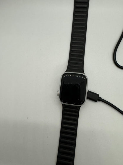 The picture shows a smartwatch with a black strap. The strap has a ridged texture. The watch face is rectangular with rounded corners and appears to be black. The watch is lying on a light-colored surface. There is also a black cable next to the watch, which seems to be a charger with a connector that likely attaches to the watch for charging.