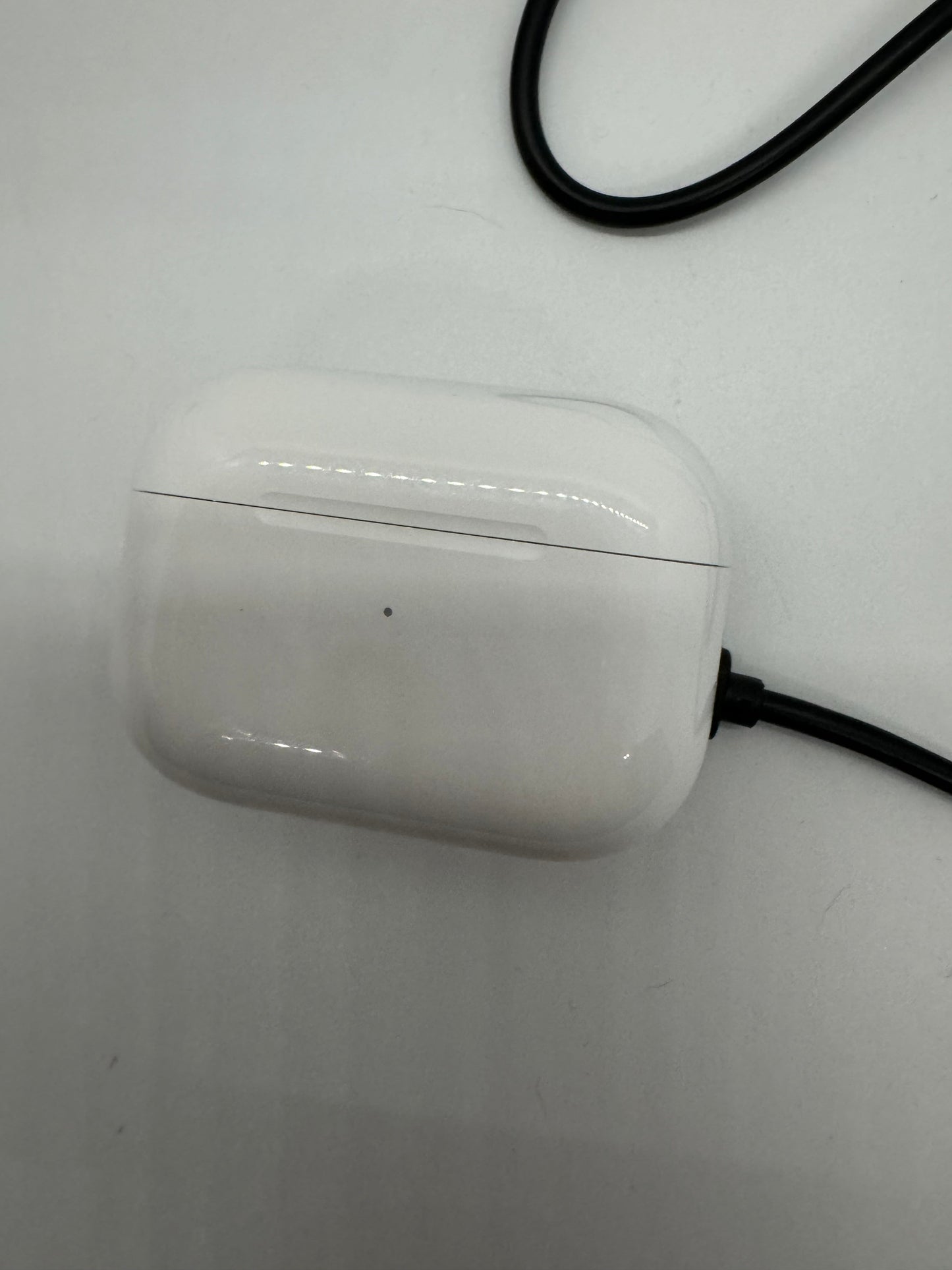 The picture shows a white AirPods case on a white surface. The case is rectangular with rounded edges and has a small LED light on the front which is not lit. There is a black cable plugged into the bottom of the case. The cable is partially visible in the top left corner of the picture. The surface the case is on appears to be a table or desk with a few minor scratches or scuffs.