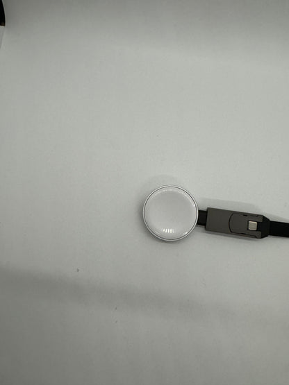 The picture shows a white surface with two objects in the bottom right corner. One object is a small, round, silver-colored item with a ridged edge. The other object is a black USB cable with a silver connector. The cable is partially visible, extending out of the frame. The rest of the image is mostly the plain white surface with a slight shadow towards the bottom.