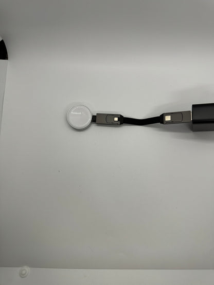 The picture shows a white surface with an object placed on it. The object appears to be a charging cable for an Apple Watch. The cable has a round magnetic charging module at one end, which is meant to attach to the back of the watch. The other end of the cable has a USB connector. The cable itself is black and flat. The magnetic charging module is silver with a white center, and the USB connector is also silver. The background is plain and white.