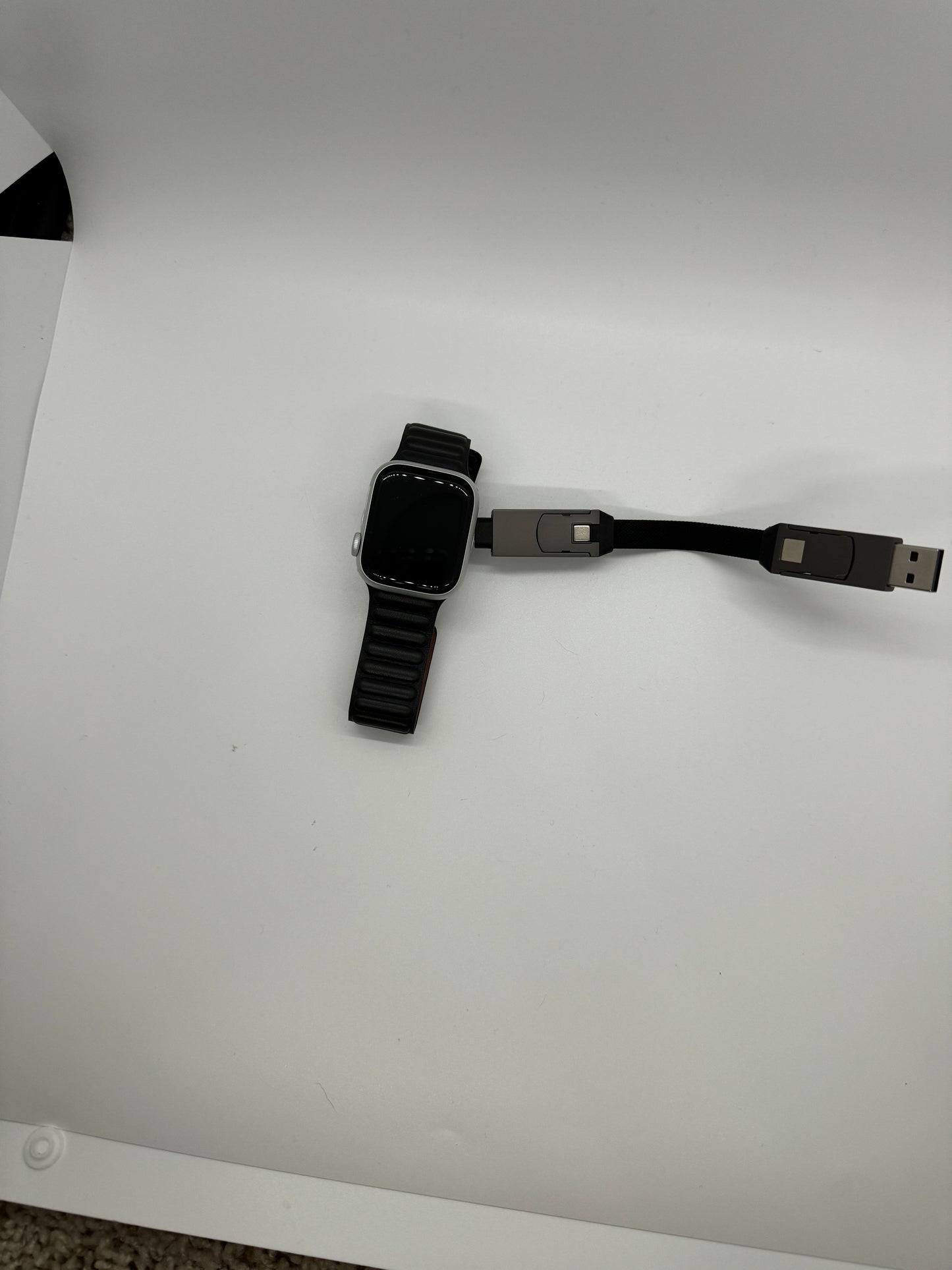Be My AI: The picture shows a smartwatch and a USB cable on a white surface. The smartwatch has a black band and a rectangular face with rounded corners. The screen of the smartwatch is black and it is placed on the left side of the image. The USB cable is on the right side of the image and has a black cord. The USB connectors are silver and one end of the cable is a standard USB, while the other end seems to be a special connector, possibly for the smartwatch. The background is plain white and there is a s