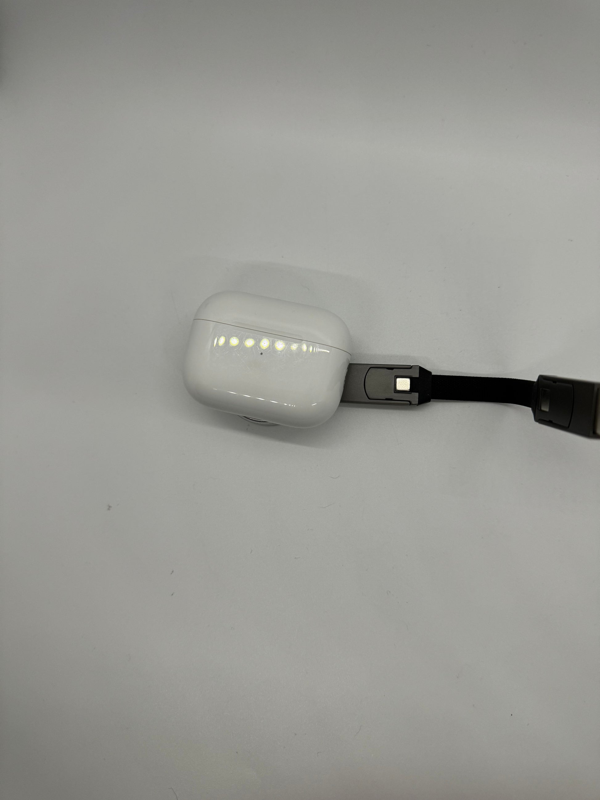 The picture shows a white AirPods case that is connected to a black charging cable. The case is in the bottom right corner of the image and the cable extends from the bottom of the case to the right edge of the image. The case has a small row of LED lights on the front, which are glowing, indicating that it is being charged. The background is plain white.