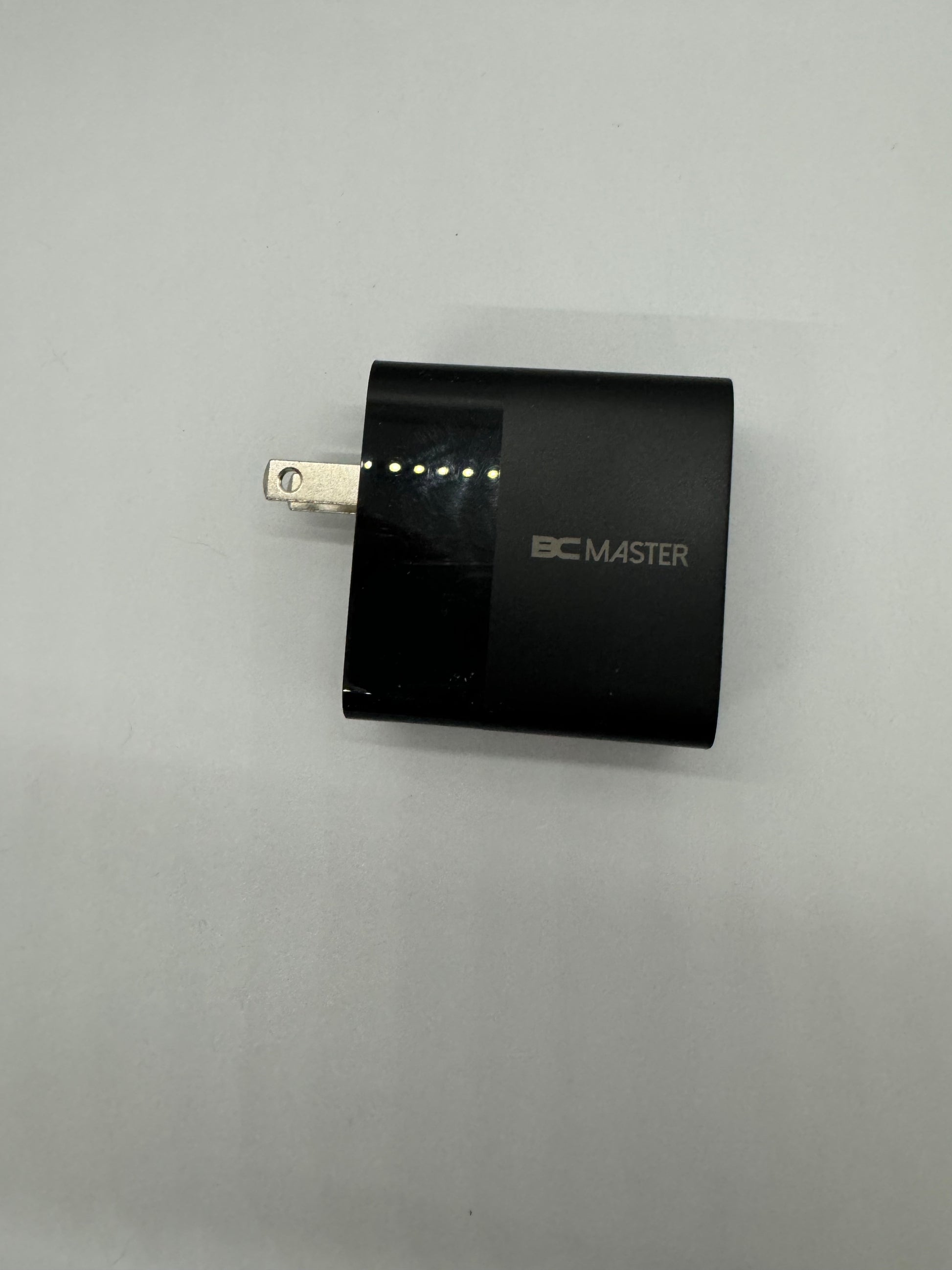 The picture shows a black rectangular adapter with a plug on one side. The plug is metallic and has two prongs for inserting into an electrical outlet. On the front side of the adapter, there is a series of small dots, possibly indicating some sort of display or indicator lights. The brand name "BC MASTER" is printed in white letters on the front side of the adapter. The background of the picture is a plain light-colored surface.