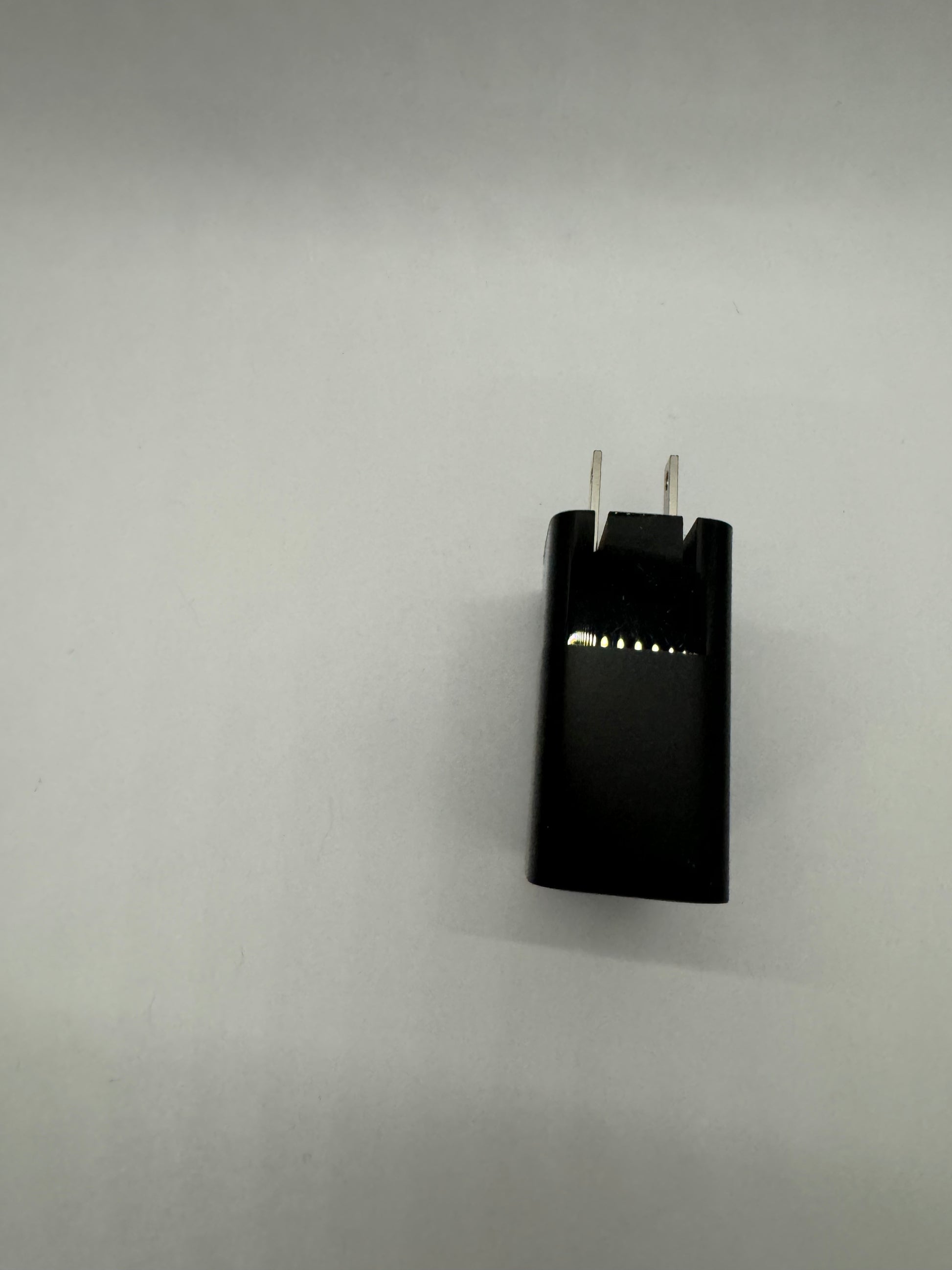 The picture shows a black rectangular object with two metal prongs sticking out from one of its shorter sides. The object appears to be a charger or adapter. The background is plain and white. The metal prongs are likely used to plug the object into an electrical outlet. There is also a series of small dots on one side of the object, which might be an indicator of some sort.