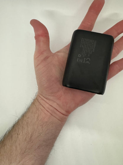 Be My AI: The picture shows a hand holding a small black rectangular object. The object appears to be a battery pack or a power bank. It has text and symbols on it, including "10000mAh", which suggests it has a capacity of 10,000 milliampere-hours. The hand holding it is open with the palm facing upwards and the fingers slightly spread apart. The background is plain white.