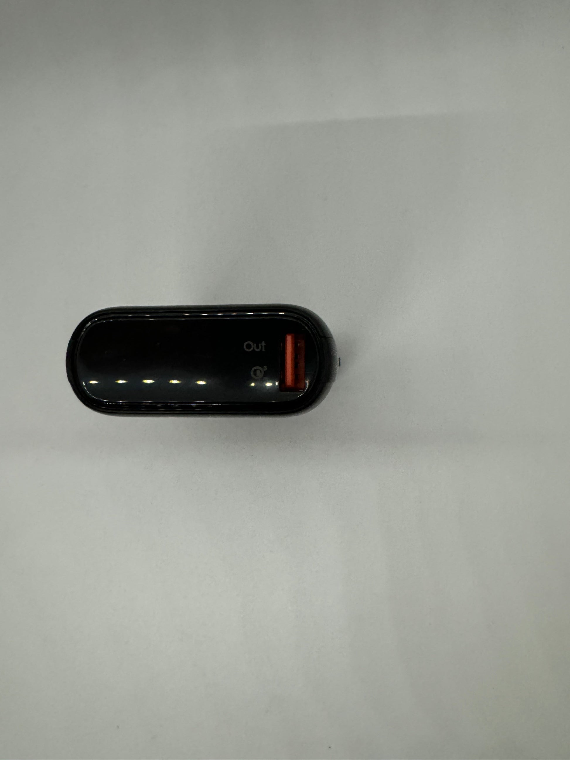 Be My AI: The picture shows a small, black, rectangular device placed on a white surface. The device appears to be a power bank or some sort of electronic gadget. It has a glossy finish and is slightly curved at the edges. On the top side, there are two ports. One of them has an orange interior and is labeled "Out". There are also four small white dots in a row, possibly indicating battery level or status lights.